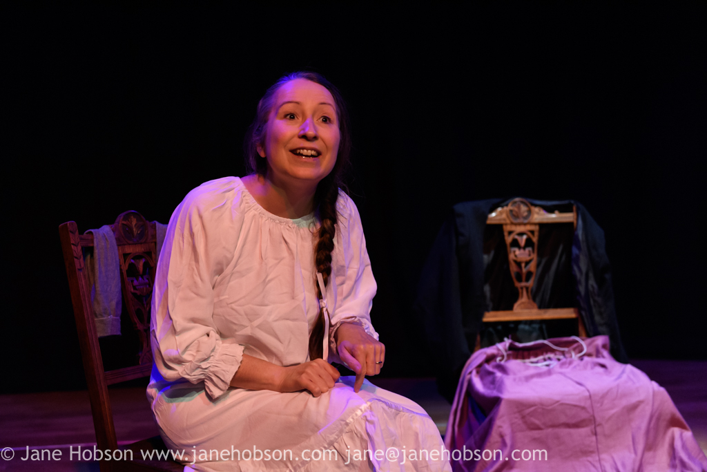 A photograph of a white actress in a white gown sitting on a chair. There is an empty chair next to her with clothes draped on it. She is addressing the audience